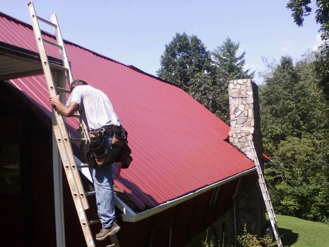 Roofer on a ladder beside a red metal roof and stone chimney — Soffit & Fascia Repair by Roofing Brothers in Salemburg